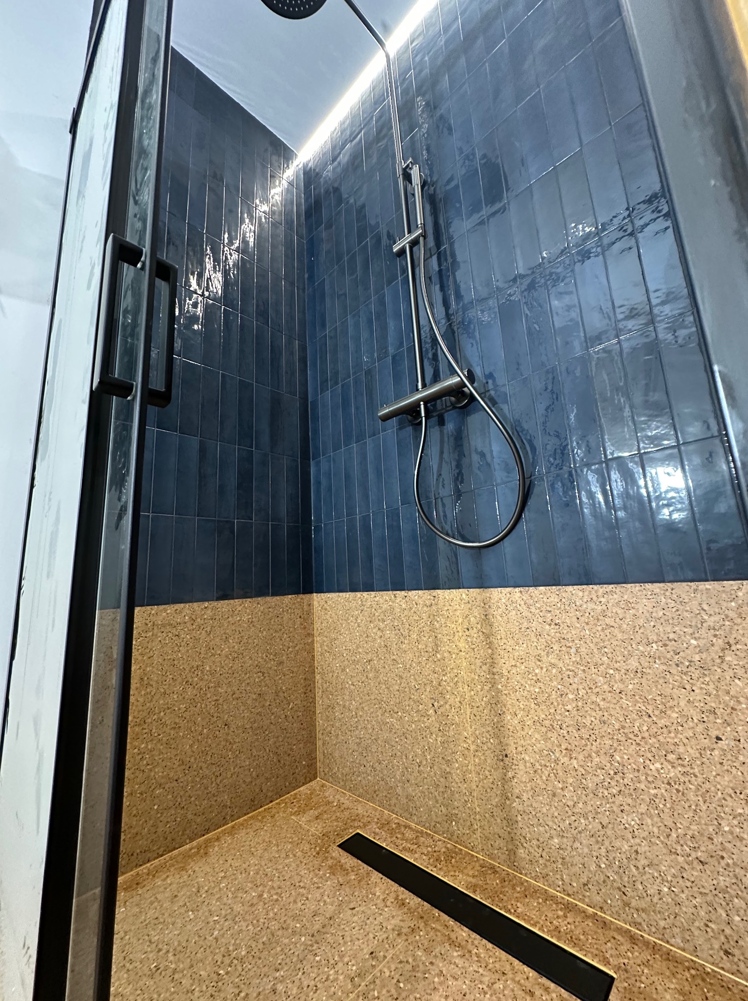 Nowoczesna kabina prysznicowa z ciemnoniebieskimi, błyszczącymi płytkami i beżową mozaiką na dole. Widoczny czarny odpływ liniowy i deszczownica.