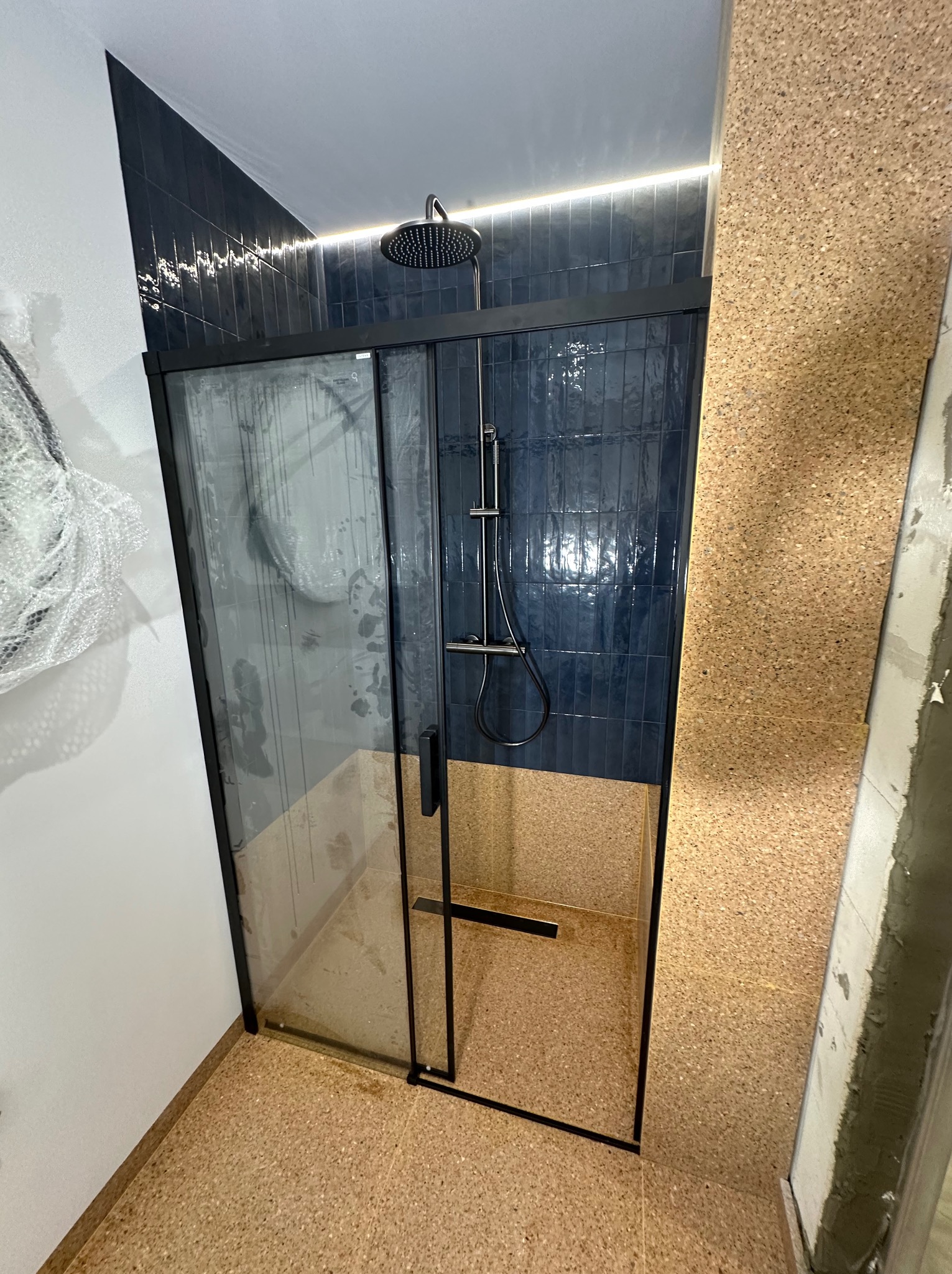 Nowoczesna kabina prysznicowa z czarną ramą i deszczownicą na tle ciemnych, błyszczących płytek ułożonych pionowo, z elementami korka na ścianach i podłodze.