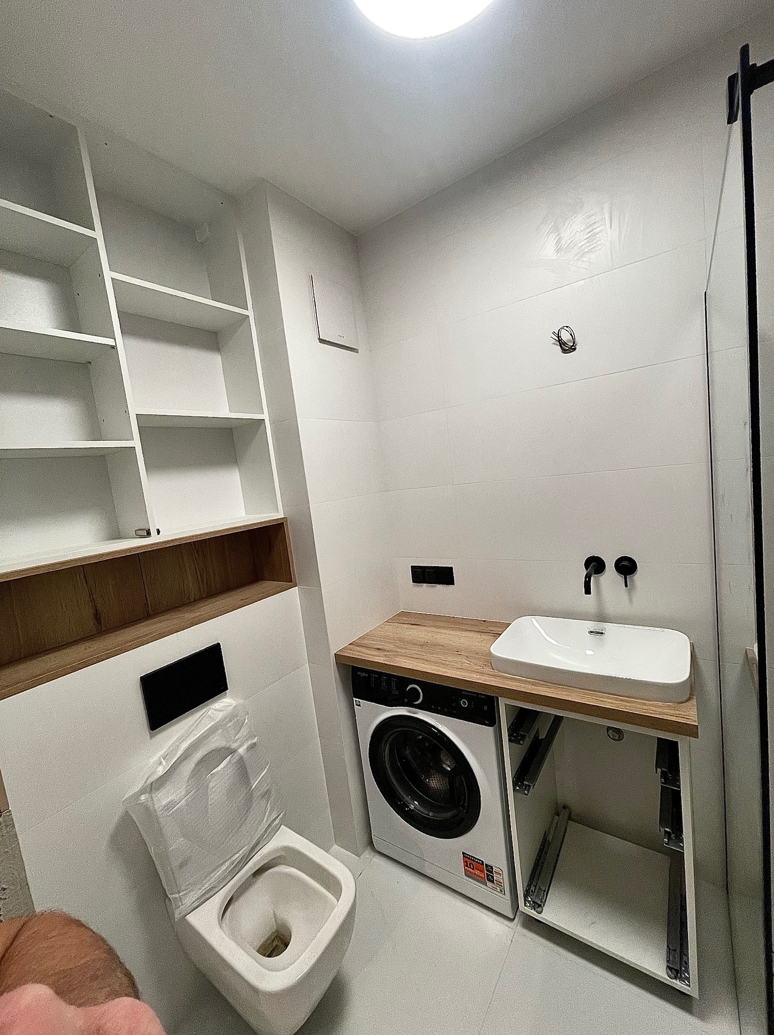 Biała łazienka po remoncie: wisząca toaleta z deską w folii, pralka pod blatem z umywalką, zabudowa meblowa z półkami. Minimalizm, nowoczesne wykończenie.