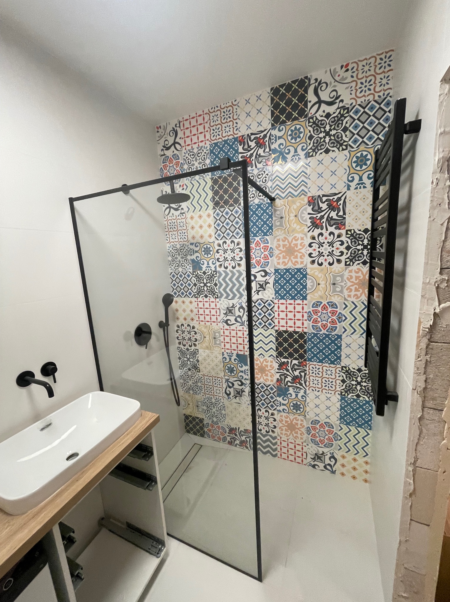 Nowoczesna łazienka z kabiną typu walk-in, czarną armaturą i ścianą wyłożoną wzorzystymi, kolorowymi płytkami. Umywalka nablatowa na szafce z drewnianym blatem.
