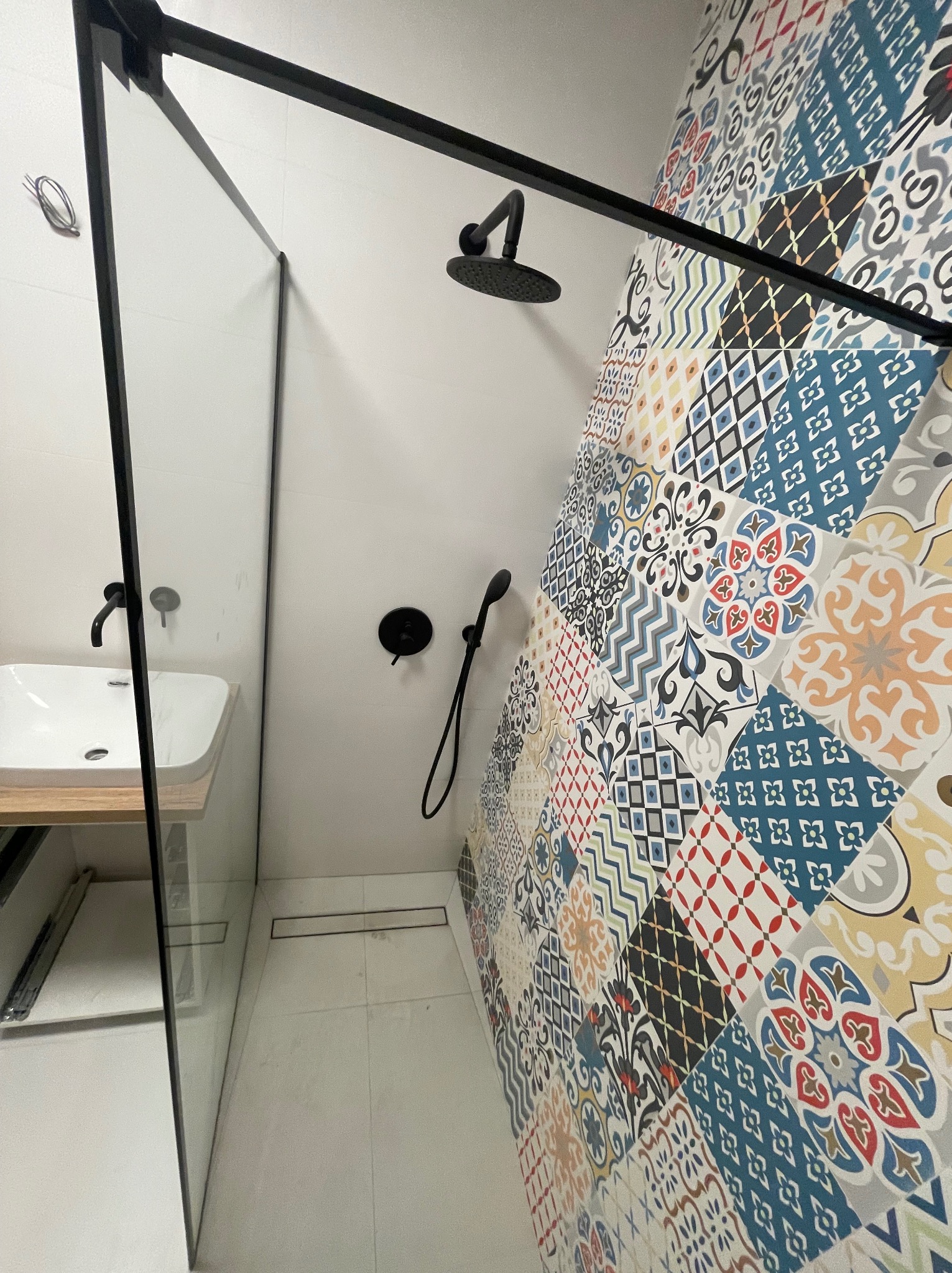 Nowoczesna łazienka z kabiną walk-in, czarną armaturą i ścianą w patchworkowe, kolorowe płytki. Umywalka nablatowa z drewnianym blatem.