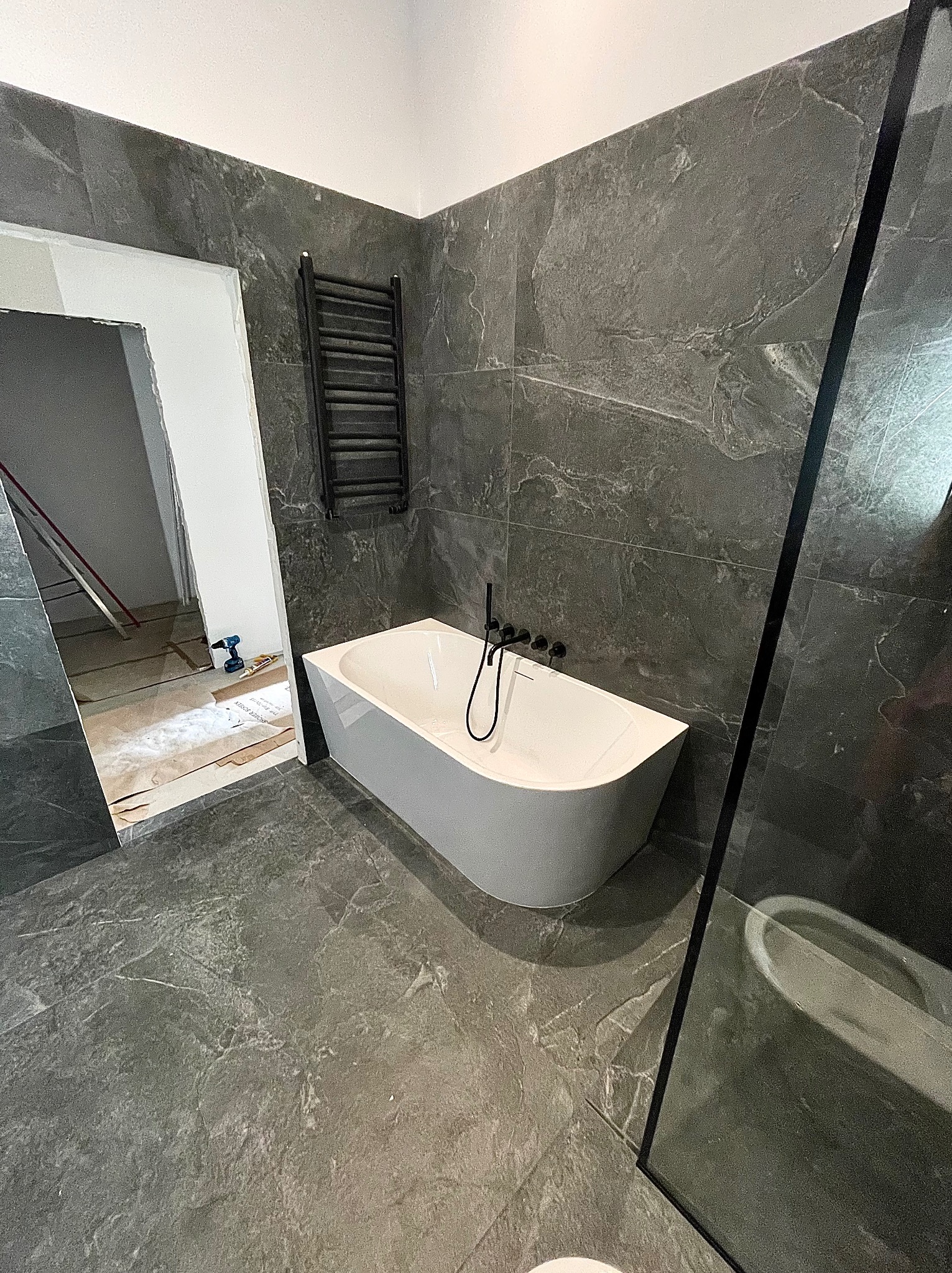 Nowoczesna łazienka z wanną wolnostojącą, grafitową armaturą i płytkami imitującymi kamień. Widoczny grzejnik drabinkowy i częściowo przeszklona kabina.