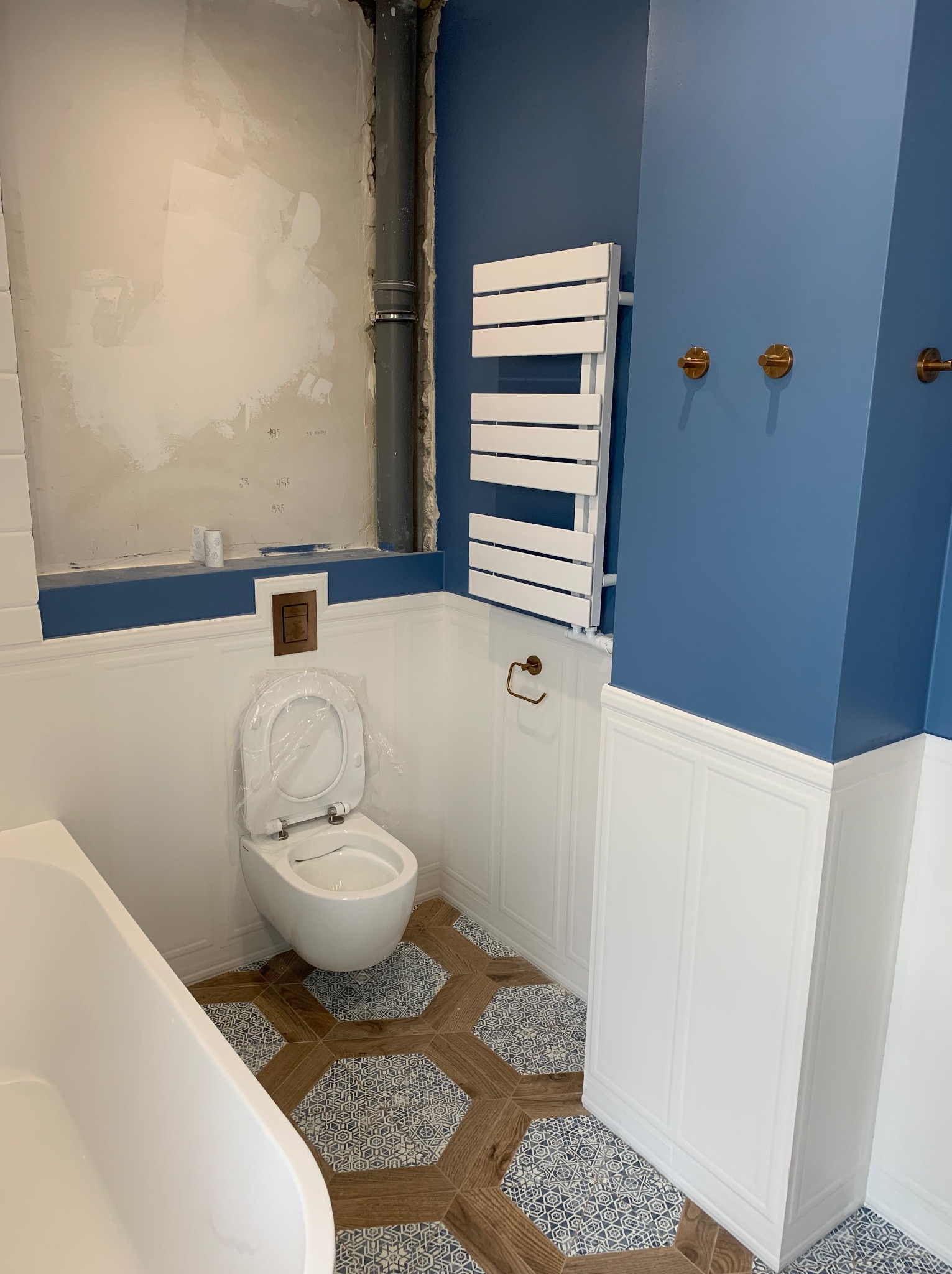 Niebiesko-biała łazienka w stylu klasycznym: mozaikowa podłoga, toaleta z deską w folii, grzejnik drabinkowy i ściana w połowie wykończona, w połowie surowa.