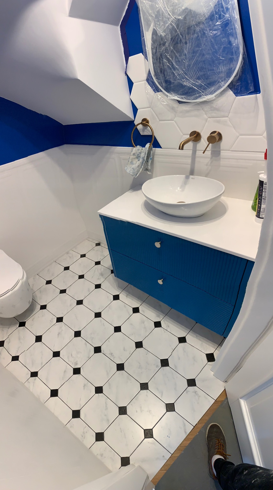 Wykończona łazienka z biało-czarną podłogą, niebieską szafką pod umywalką i lustrem zabezpieczonym folią. Widoczne złote elementy armatury i białe heksagonalne płytki.