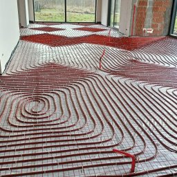 HYDROLUX MARCIN FELUKS - Rozłożona instalacja ogrzewania podłogowego z czerwonych rurek na folii, widok z góry, duże okna w tle, surowy stan budynku.