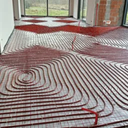 Rozłożona instalacja ogrzewania podłogowego z czerwonych rurek na folii, widok z góry, duże okna w tle, surowy stan budynku.