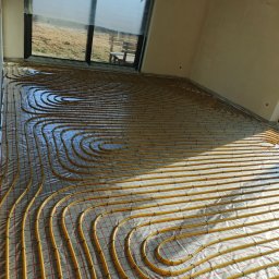 HYDROLUX MARCIN FELUKS - Żółte rury ogrzewania podłogowego ułożone na folii w surowym wnętrzu z dużym oknem. Widok na zewnątrz, gdzie widać trawę i ławkę.