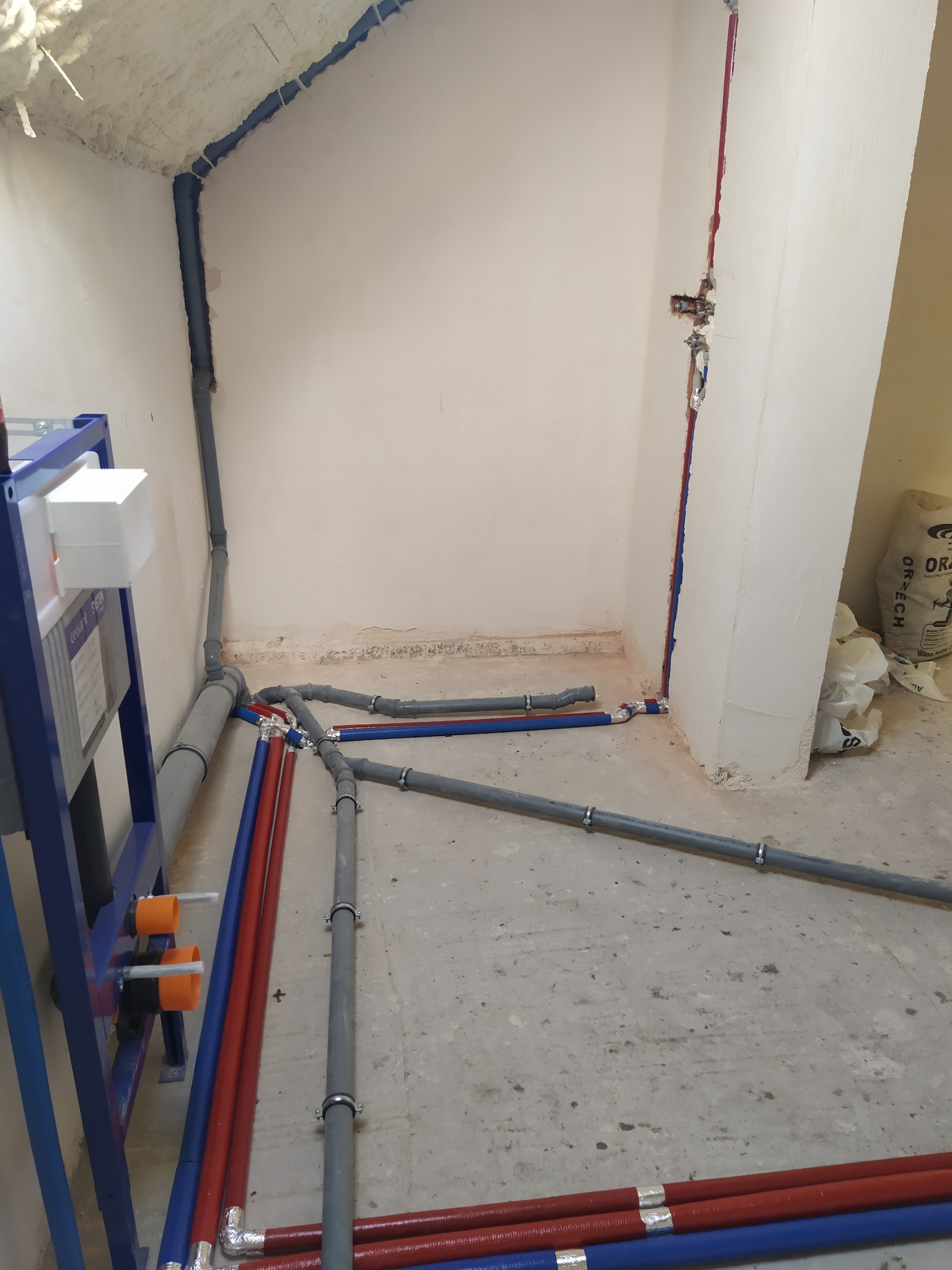 Szare i kolorowe rury instalacji hydraulicznej ułożone na surowym podłożu w trakcie montażu, widoczne elementy mocujące i fragment stelaża podtynkowego.