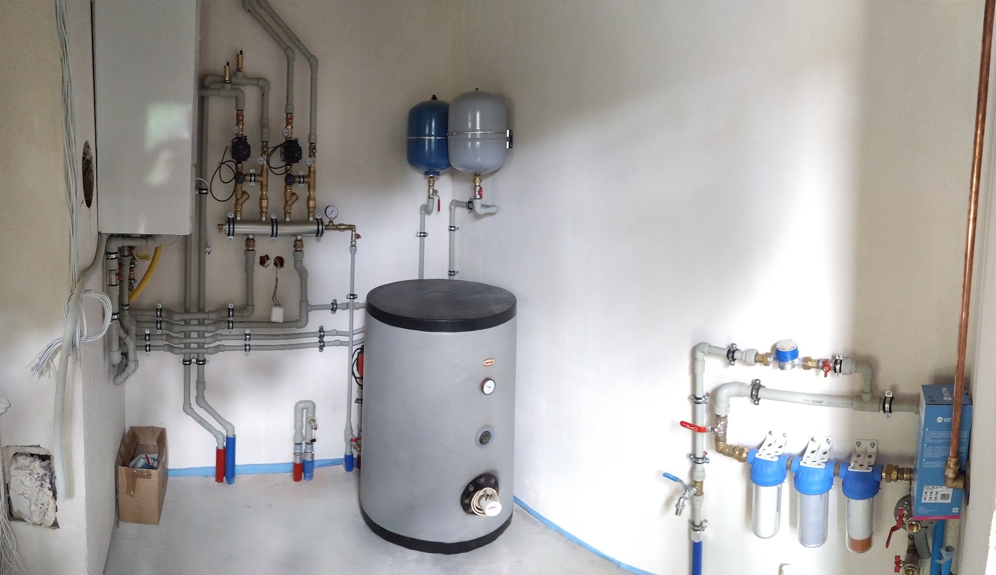 Zakończona instalacja CO z piecem, zbiornikiem, naczyniami wzbiorczymi i filtrami wody. Widoczne rury z szarego PCV i miedziane. Białe ściany, surowy stan wykończenia.