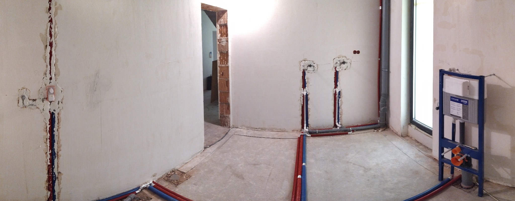 Wnętrze w trakcie prac hydraulicznych: widoczne rury (czerwone, niebieskie, szare) w ścianach i na podłodze, stelaż podtynkowy WC przy oknie, surowe ściany.