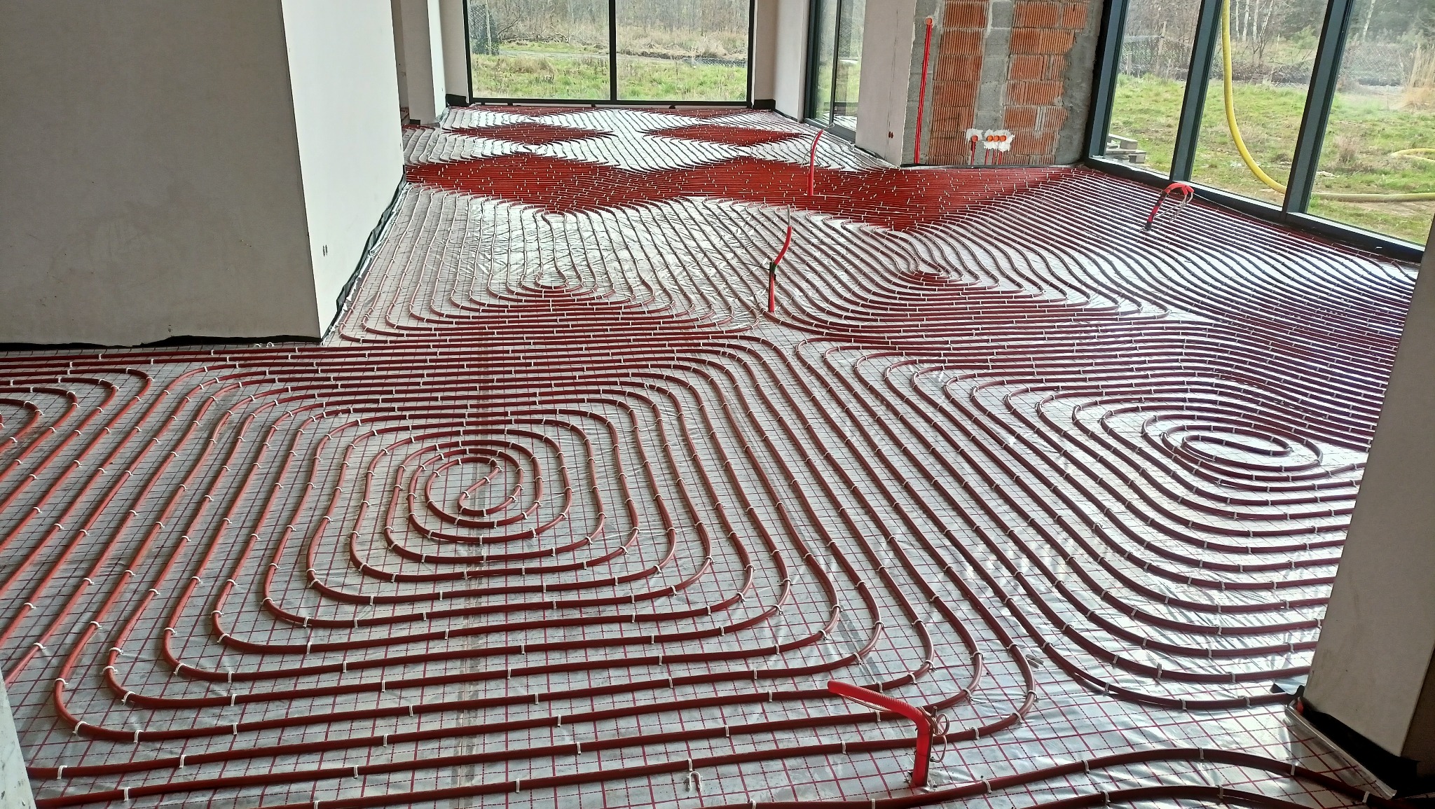 Rozłożona instalacja ogrzewania podłogowego z czerwonych rurek na folii, widok z góry, duże okna w tle, surowy stan budynku.