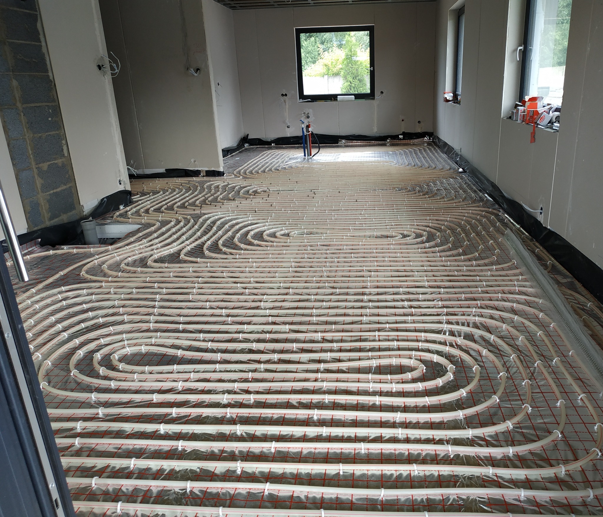 Rozłożona instalacja ogrzewania podłogowego w nowym budynku, widoczne rury na folii z siatką, okna w tle, surowy stan.
