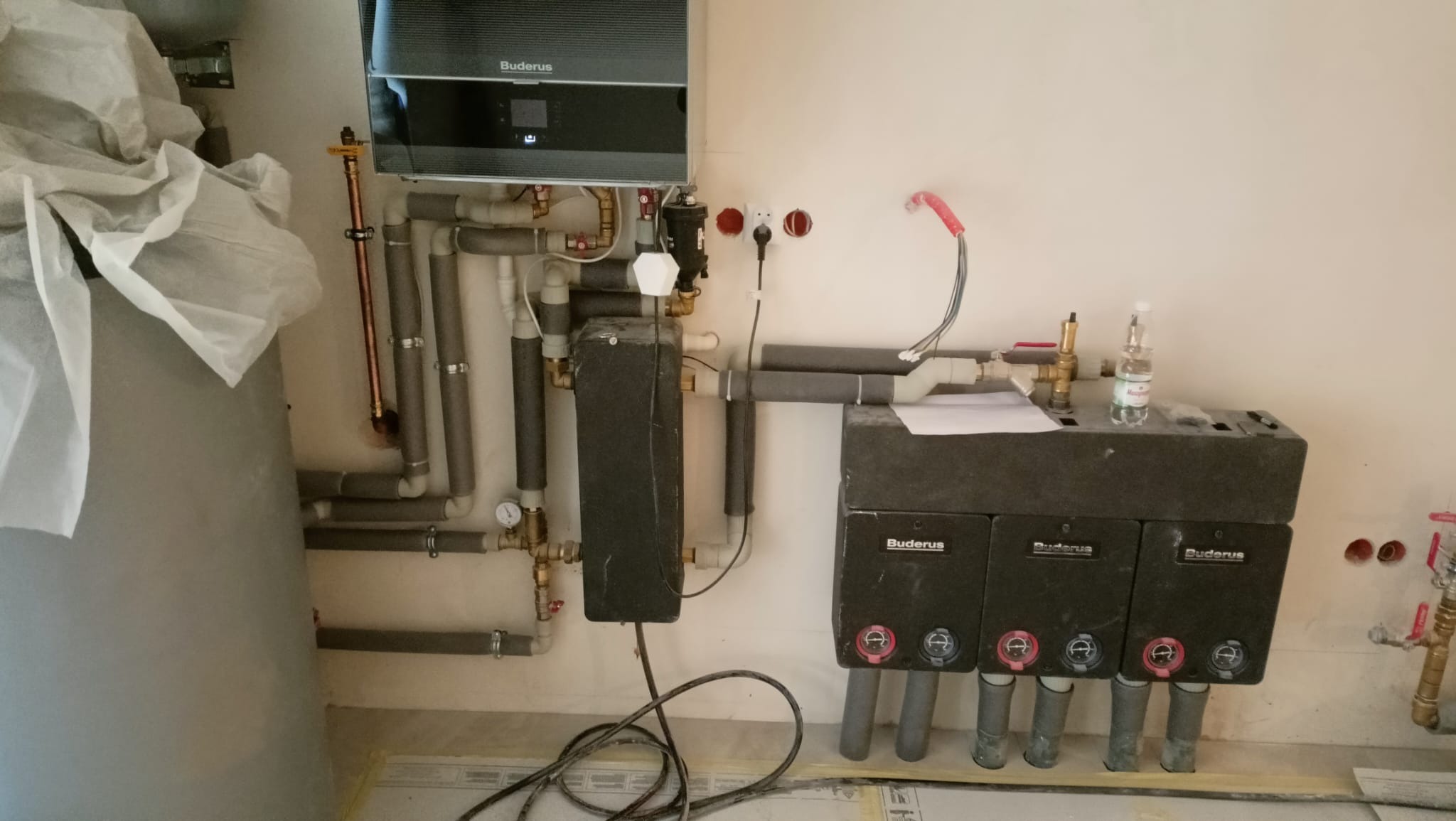 Kompleksowa instalacja grzewcza Buderus: piec, zasobnik, rozdzielacze i rury w szarej izolacji. Widoczne przyłącza i elementy montażowe. Prace w trakcie realizacji.