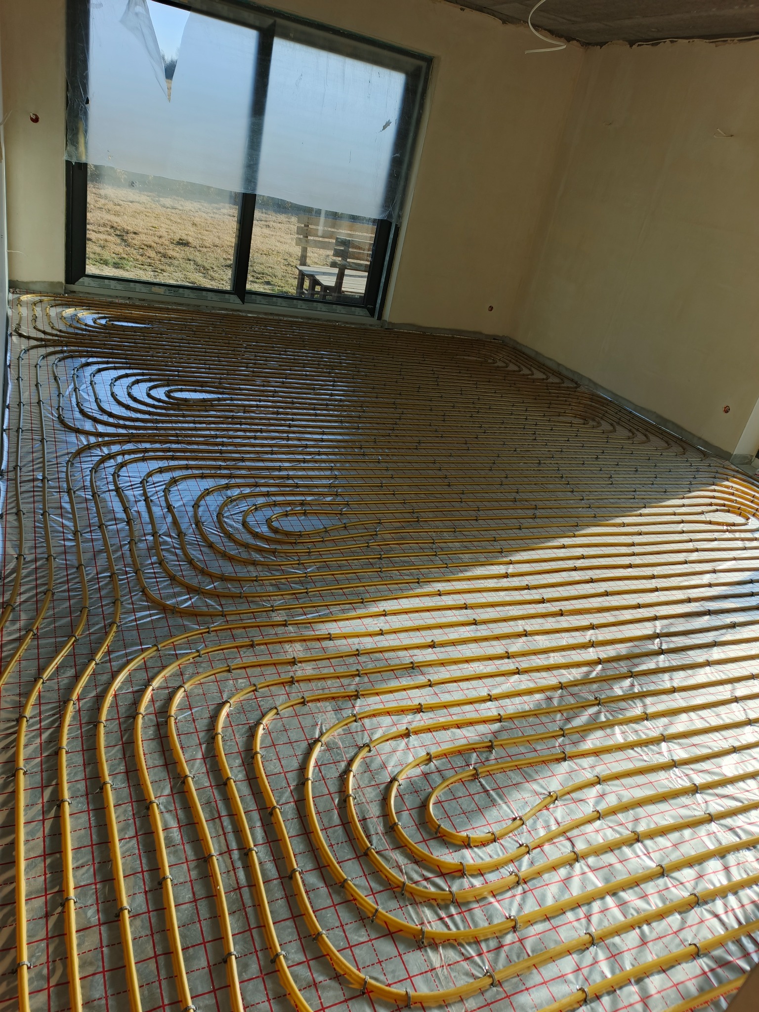 Żółte rury ogrzewania podłogowego ułożone na folii w surowym wnętrzu z dużym oknem. Widok na zewnątrz, gdzie widać trawę i ławkę.