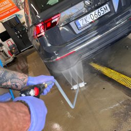 RENT-IPSUM TOMASZ ZIENTARA - Szary Volkswagen Tiguan myty pod ciśnieniem w myjni. Woda spływa po karoserii w kierunku żółtej kratki odpływowej. Widoczne wytatuowane ramię pracownika w rękawicy.