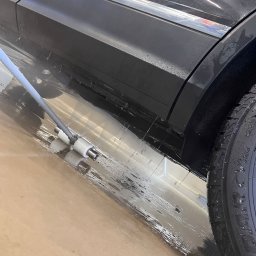 RENT-IPSUM TOMASZ ZIENTARA - Czarny samochód w trakcie mycia podwozia specjalistycznym urządzeniem z dyszami, widoczne krople wody i mokra posadzka. Opona z bliska.