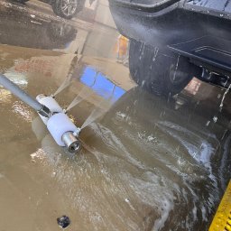 RENT-IPSUM TOMASZ ZIENTARA - Czarny samochód poddawany myciu podwozia w myjni automatycznej. Woda pod ciśnieniem spryskuje podwozie pojazdu, tworząc efektowną fontannę.