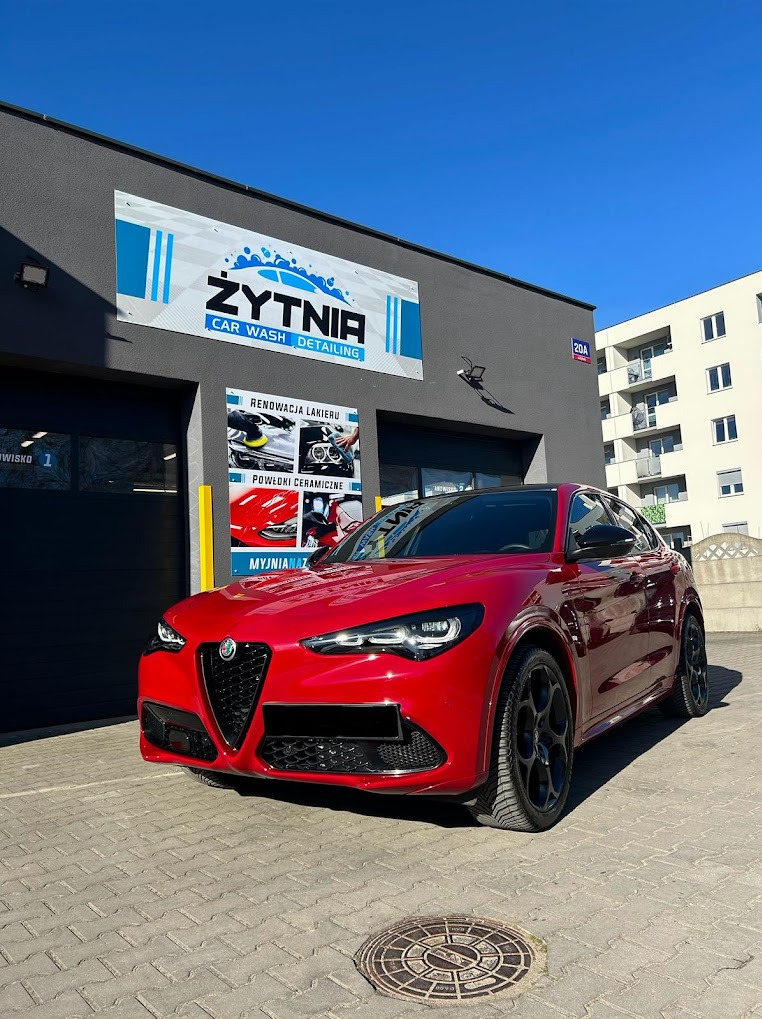 Czerwona Alfa Romeo Stelvio przed budynkiem myjni detailingowej z szyldem 'Żytnia Car Wash Detailing', w słoneczny dzień. Widoczny fragment chodnika z włazem.