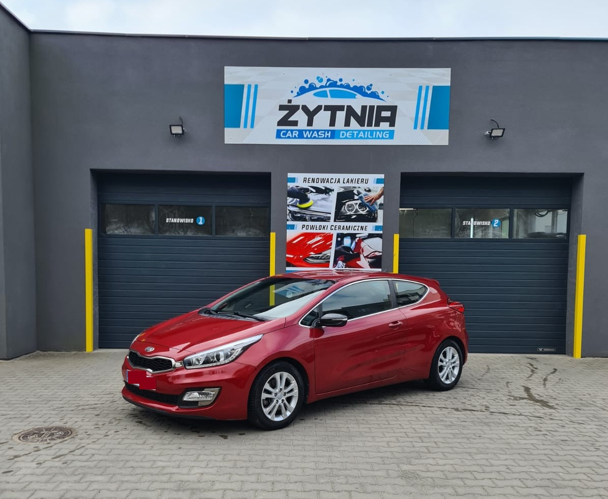 Czerwone auto przed myjnią Żytnia Car Wash Detailing z szyldem i plakatami o renowacji lakieru i powłokach ceramicznych. Widoczne stanowiska myjni.