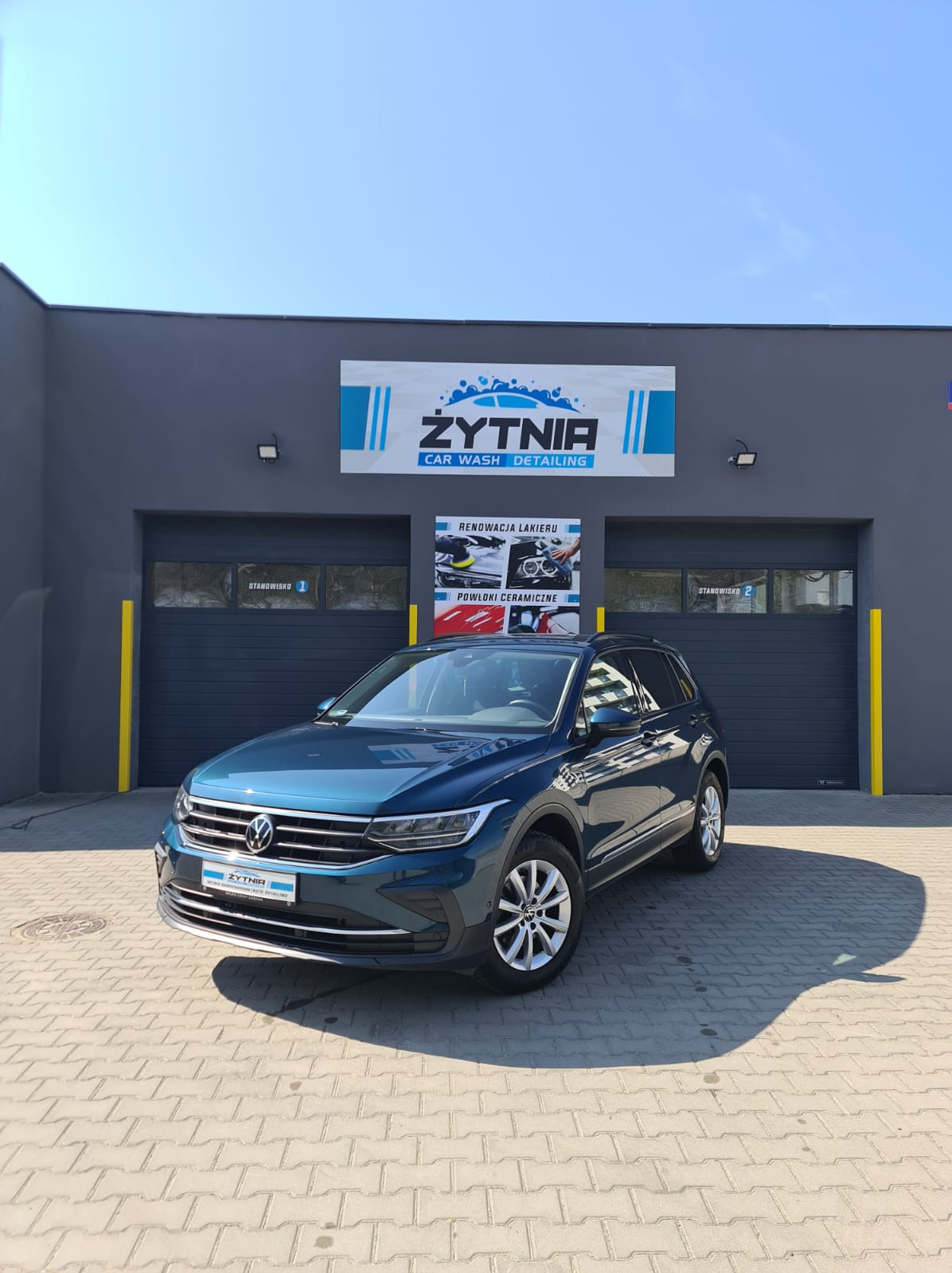 Niebieski Volkswagen Tiguan po myciu, zaparkowany przed myjnią Żytnia Car Wash Detailing z szarym budynkiem i żółtymi akcentami. Słoneczny dzień.