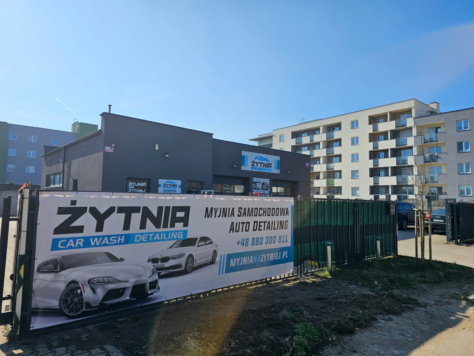 Budynek myjni samochodowej Żytnia z banerem reklamowym prezentującym sportowe i luksusowe auta. W tle budynek mieszkalny i błękitne niebo.