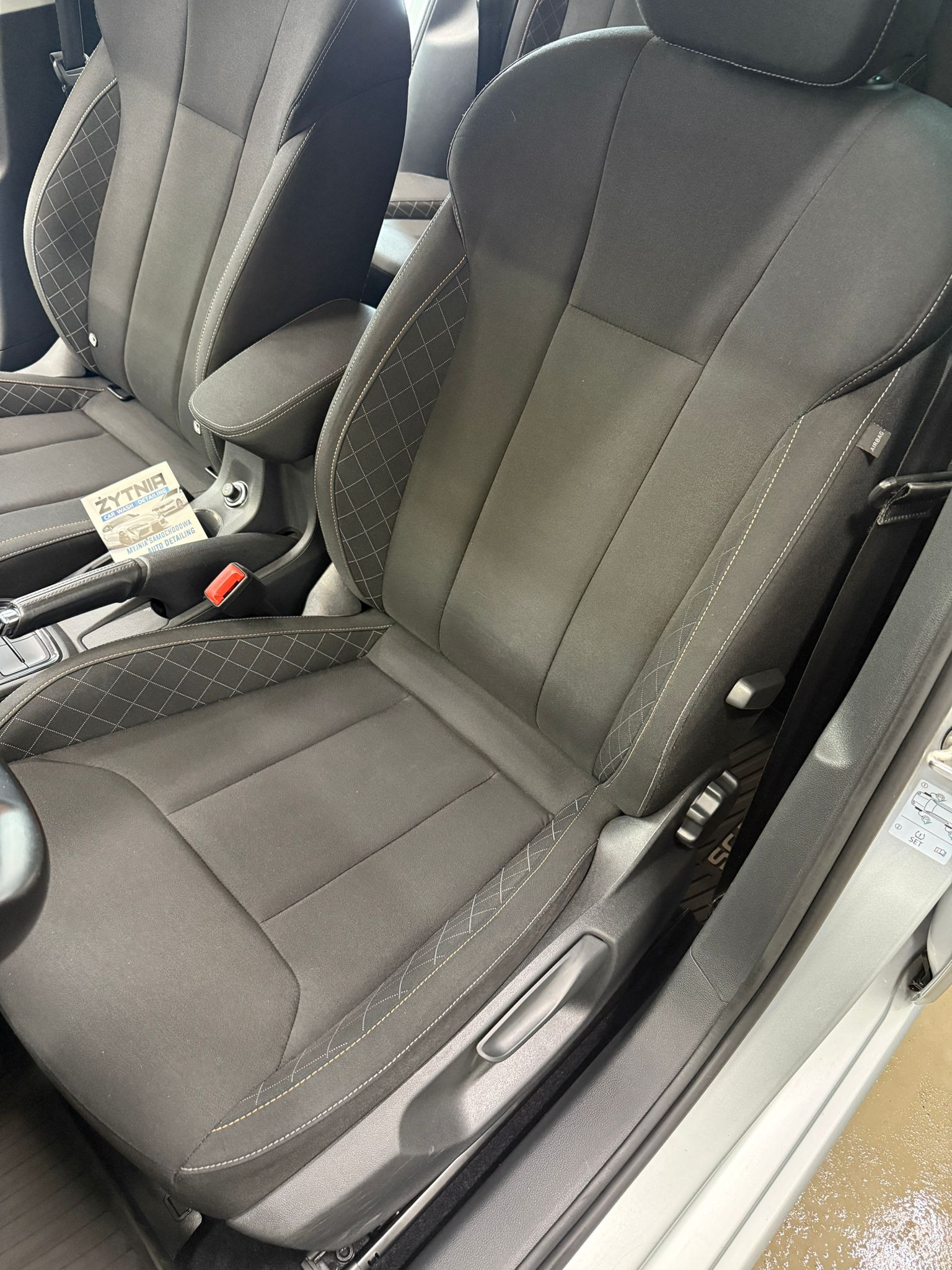 Ciemna tapicerka samochodowa z jasnymi przeszyciami w romby, widoczny fragment deski rozdzielczej i ulotka reklamowa myjni.