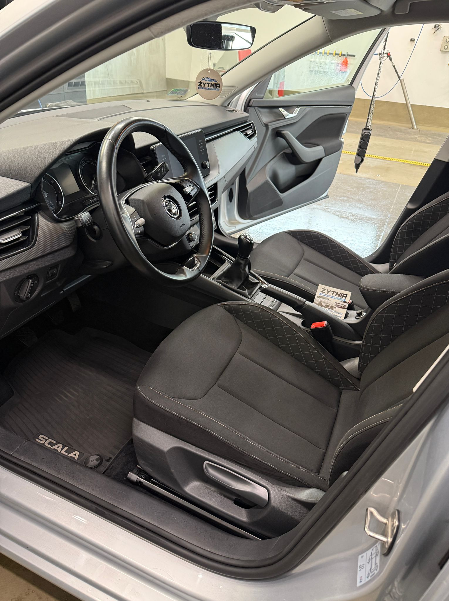 Wnętrze srebrnej Skody Scala po detailingu: czyste fotele z jasnymi przeszyciami, gumowe dywaniki z logo, otwarte drzwi, zawieszka zapachowa.