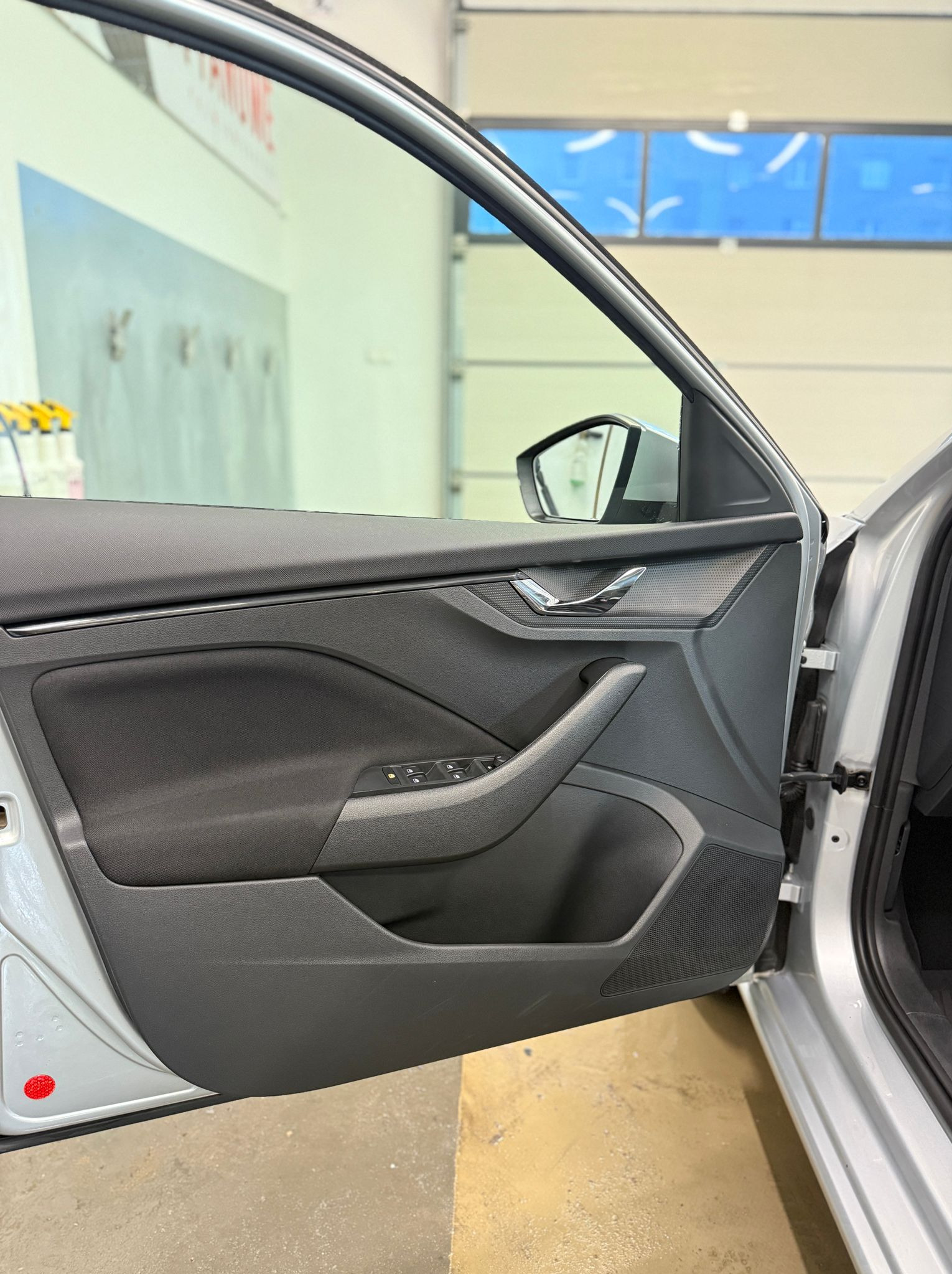 Otwarta, przednia, srebrna  drzwi samochodu Skoda z widocznym panelem sterowania szybami i lusterkiem bocznym w tle, na tle wnętrza myjni.