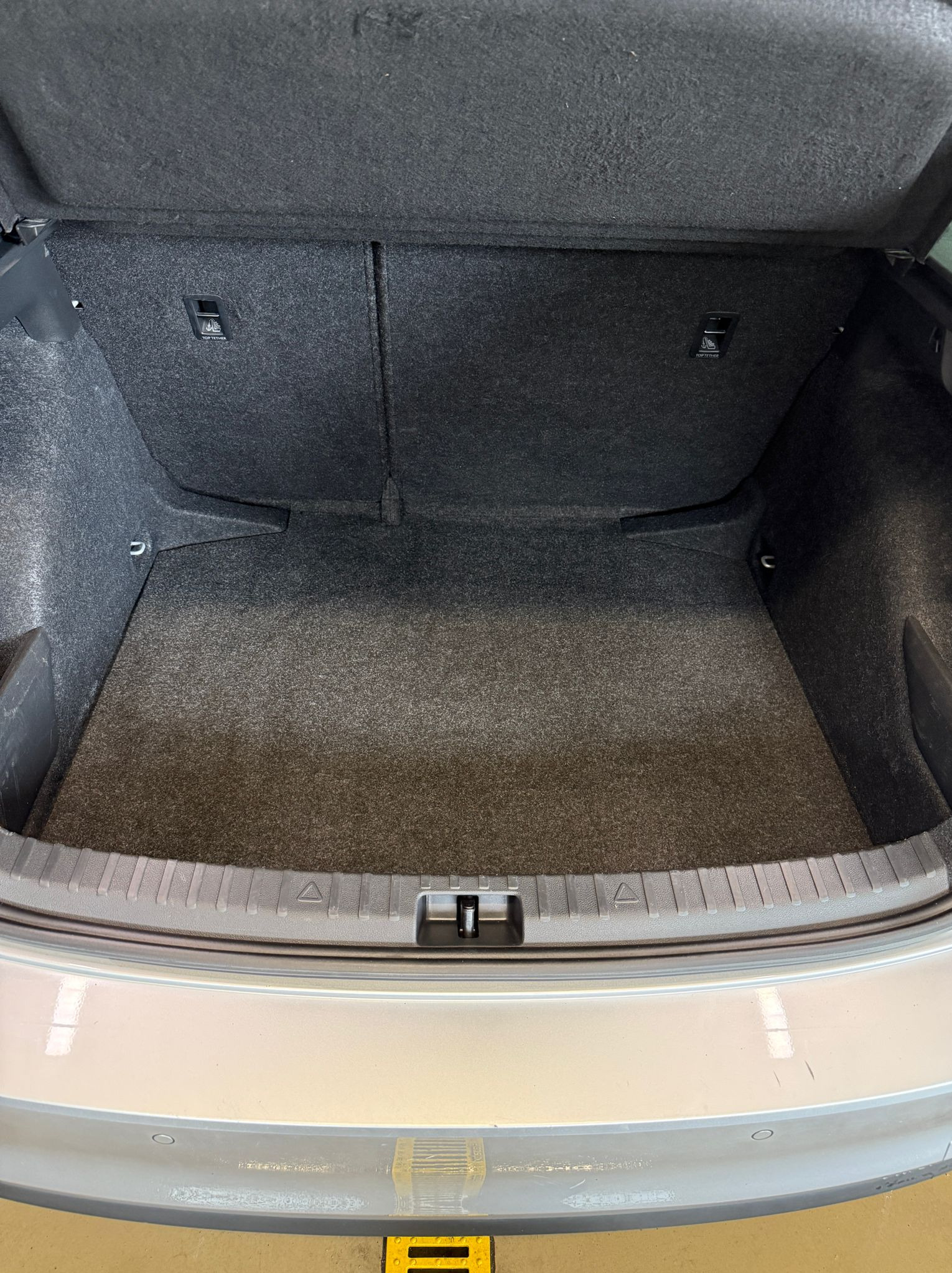 Wnętrze bagażnika samochodu osobowego z szarą tapicerką i widocznymi uchwytami mocującymi. Widok z góry na pustą przestrzeń bagażową.