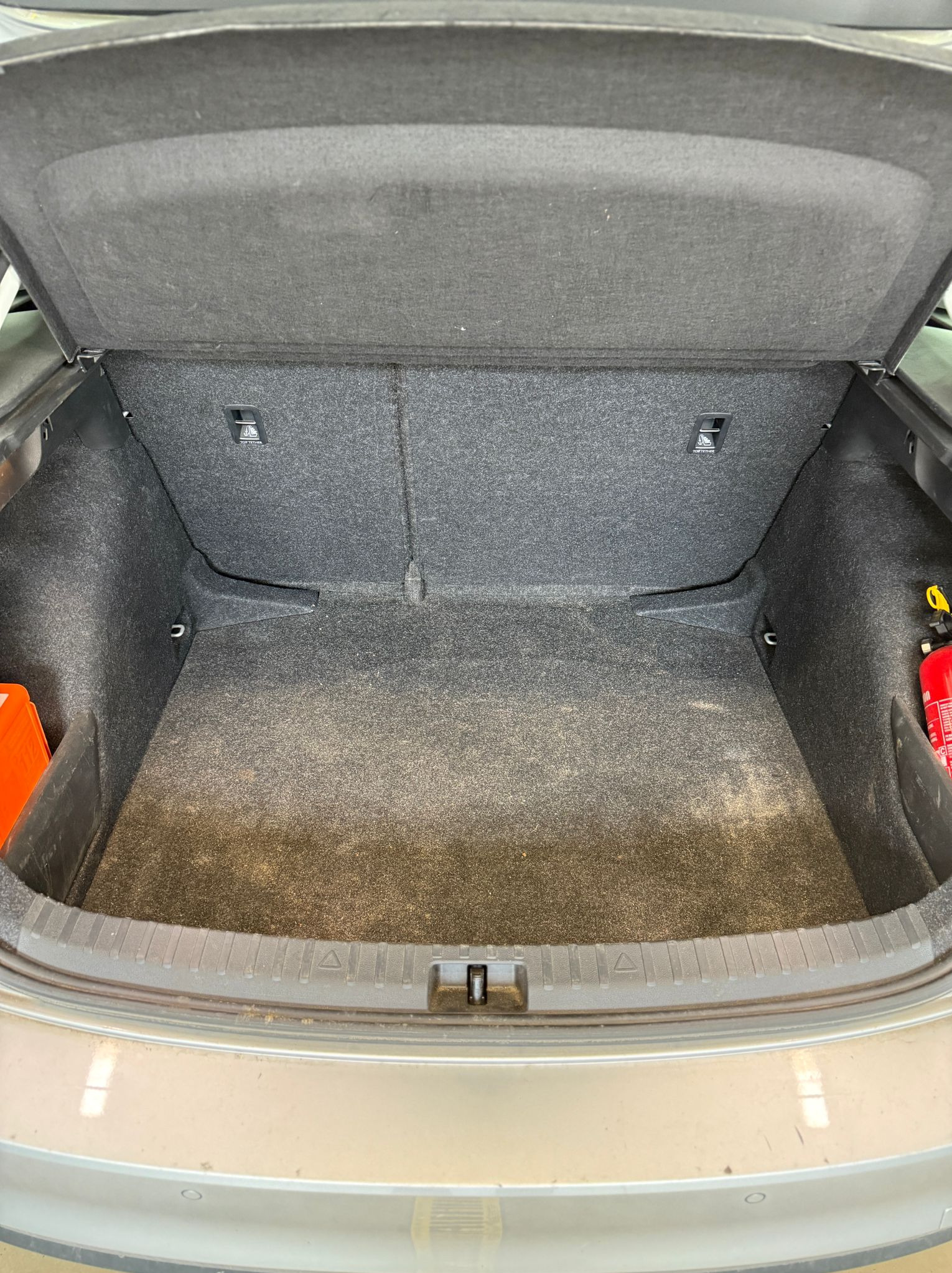 Wnętrze bagażnika samochodu z otwartą klapą, widoczna szara tapicerka i elementy wyposażenia. W tle pomarańczowy pojemnik i gaśnica.