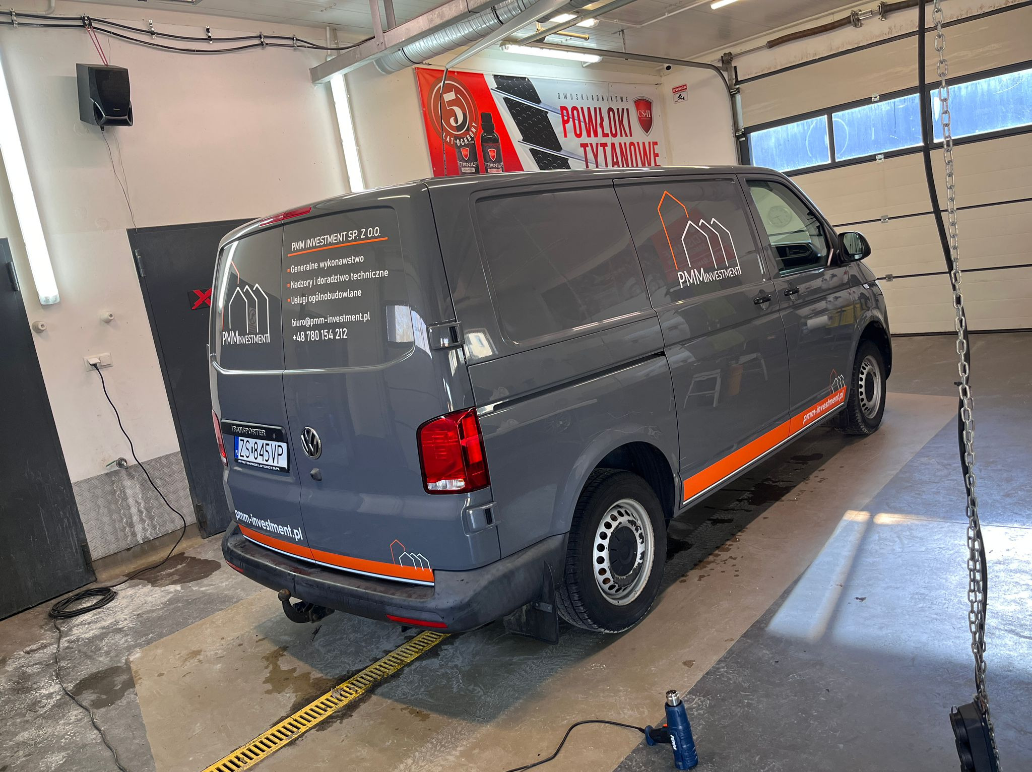 Szary van z logo PMM Investment i pomarańczowymi akcentami, zaparkowany w warsztacie z banerem reklamowym powłok tytanowych w tle. Widoczny opalarka.