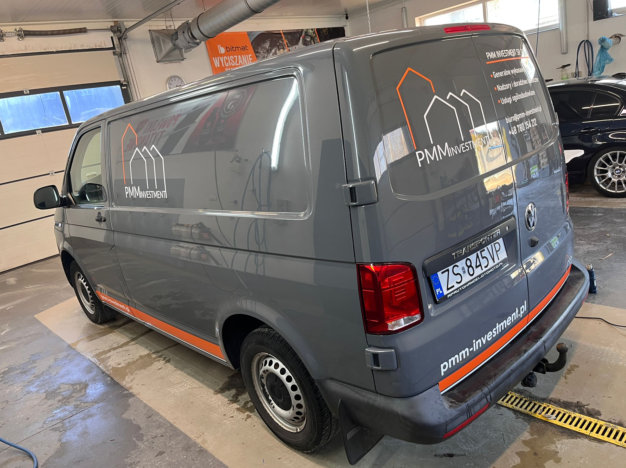 Szary van z pomarańczowymi akcentami i logo PMM Investment, zaparkowany w myjni. Widoczne dane kontaktowe firmy na karoserii. Świeżo umyty, mokra podłoga.