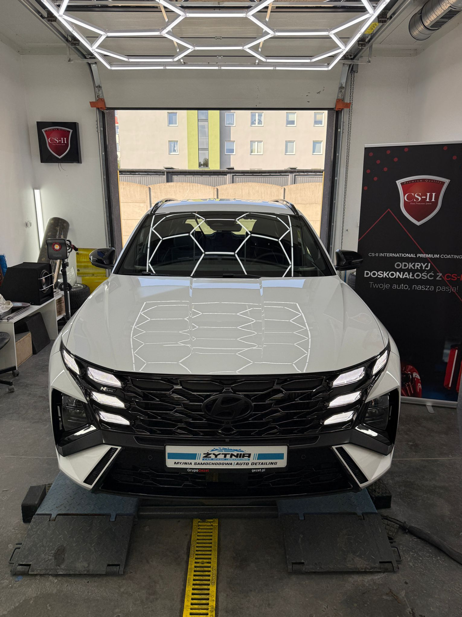 Biały SUV Hyundai Tucson po detailingu, z widocznymi odbiciami heksagonalnych lamp na karoserii, stojący w jasnym garażu z logo CS-II w tle.