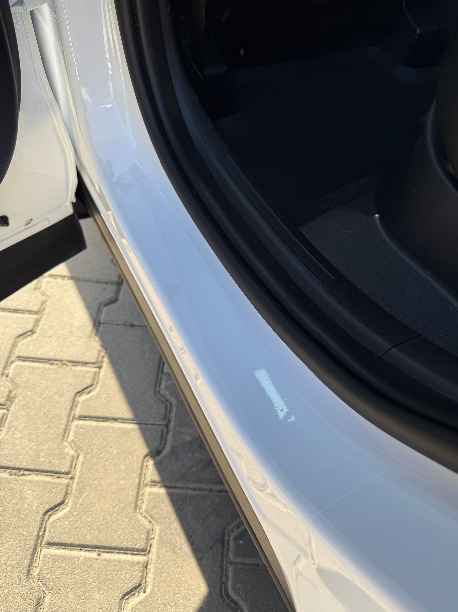 Błyszczący, biały próg samochodu po detailingu, z widocznym odbiciem światła i czarnym uszczelnieniem drzwi. Kostka brukowa w tle.