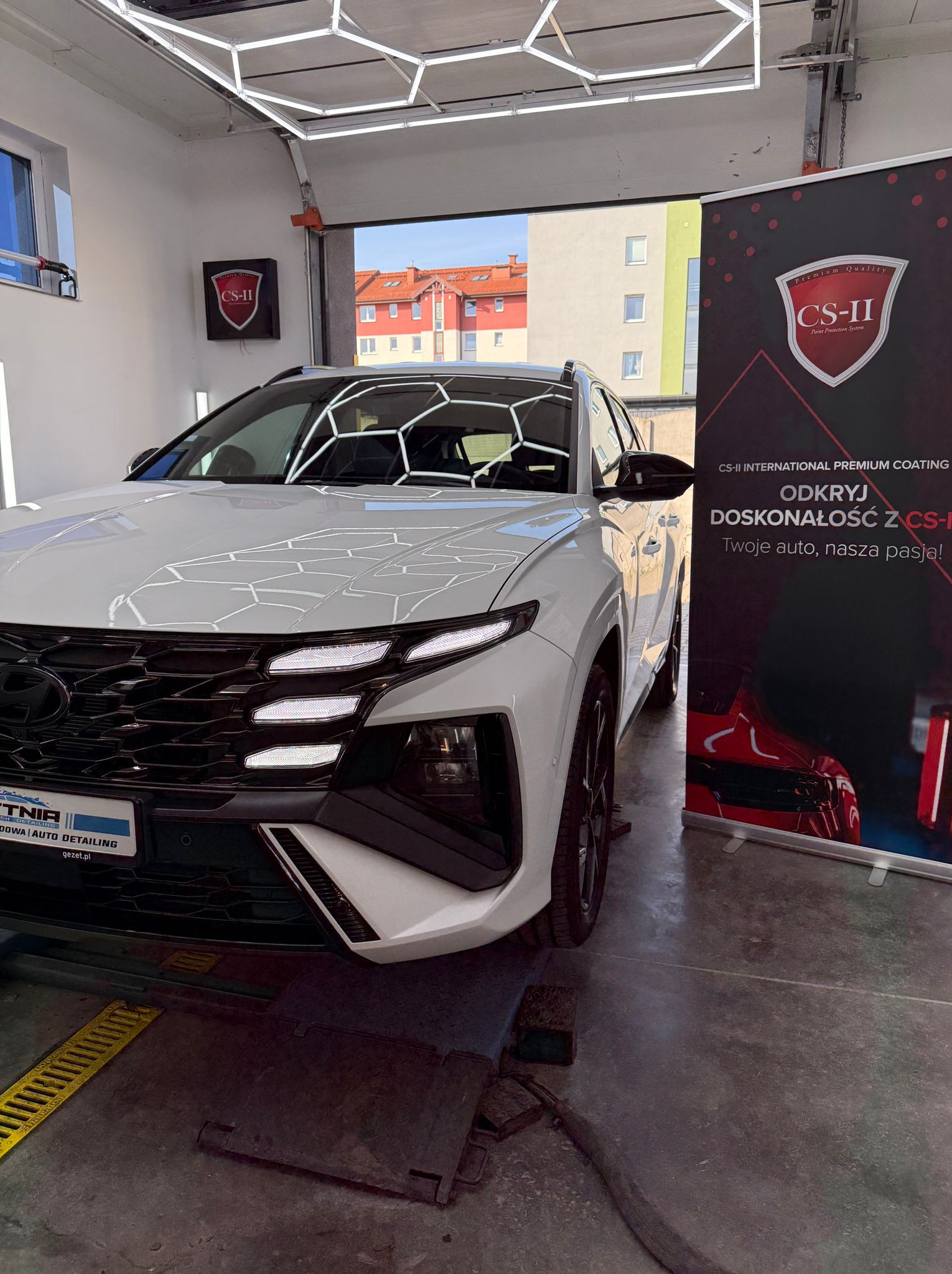 Biały Hyundai Tucson po detailingu w studiu, z widocznym logo CS-II i oświetleniem heksagonalnym. Auto lśni, podkreślając profesjonalizm usługi.