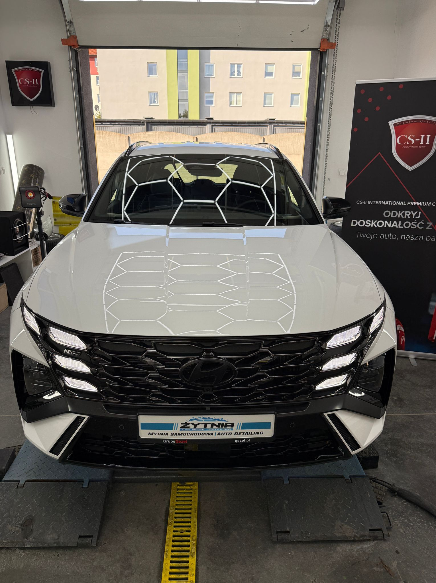 Biały Hyundai Tucson po detailingu w myjni Żytnia, z widocznym odbiciem heksagonalnej struktury na masce. W tle brama garażowa i budynek.