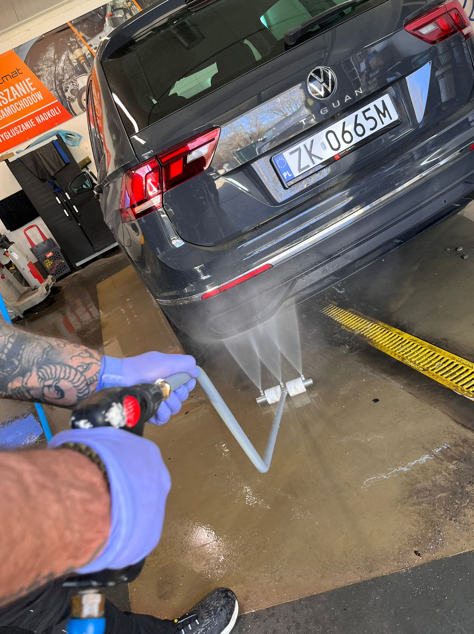 Szary Volkswagen Tiguan myty pod ciśnieniem w myjni. Woda spływa po karoserii w kierunku żółtej kratki odpływowej. Widoczne wytatuowane ramię pracownika w rękawicy.