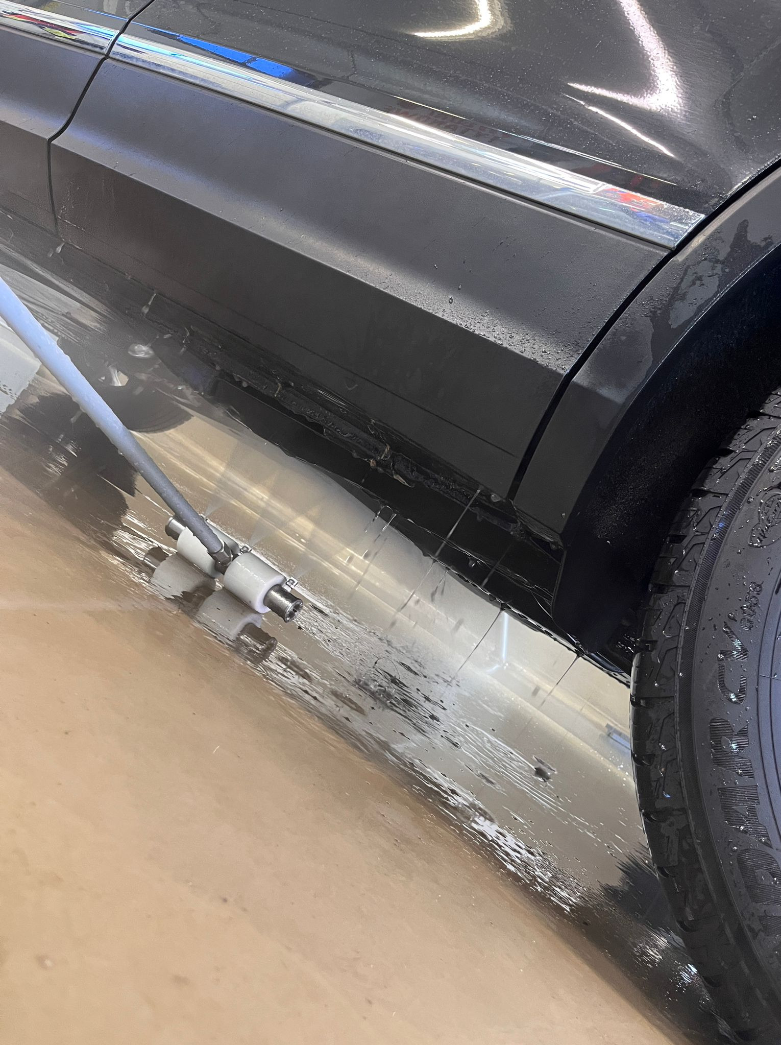 Czarny samochód w trakcie mycia podwozia specjalistycznym urządzeniem z dyszami, widoczne krople wody i mokra posadzka. Opona z bliska.