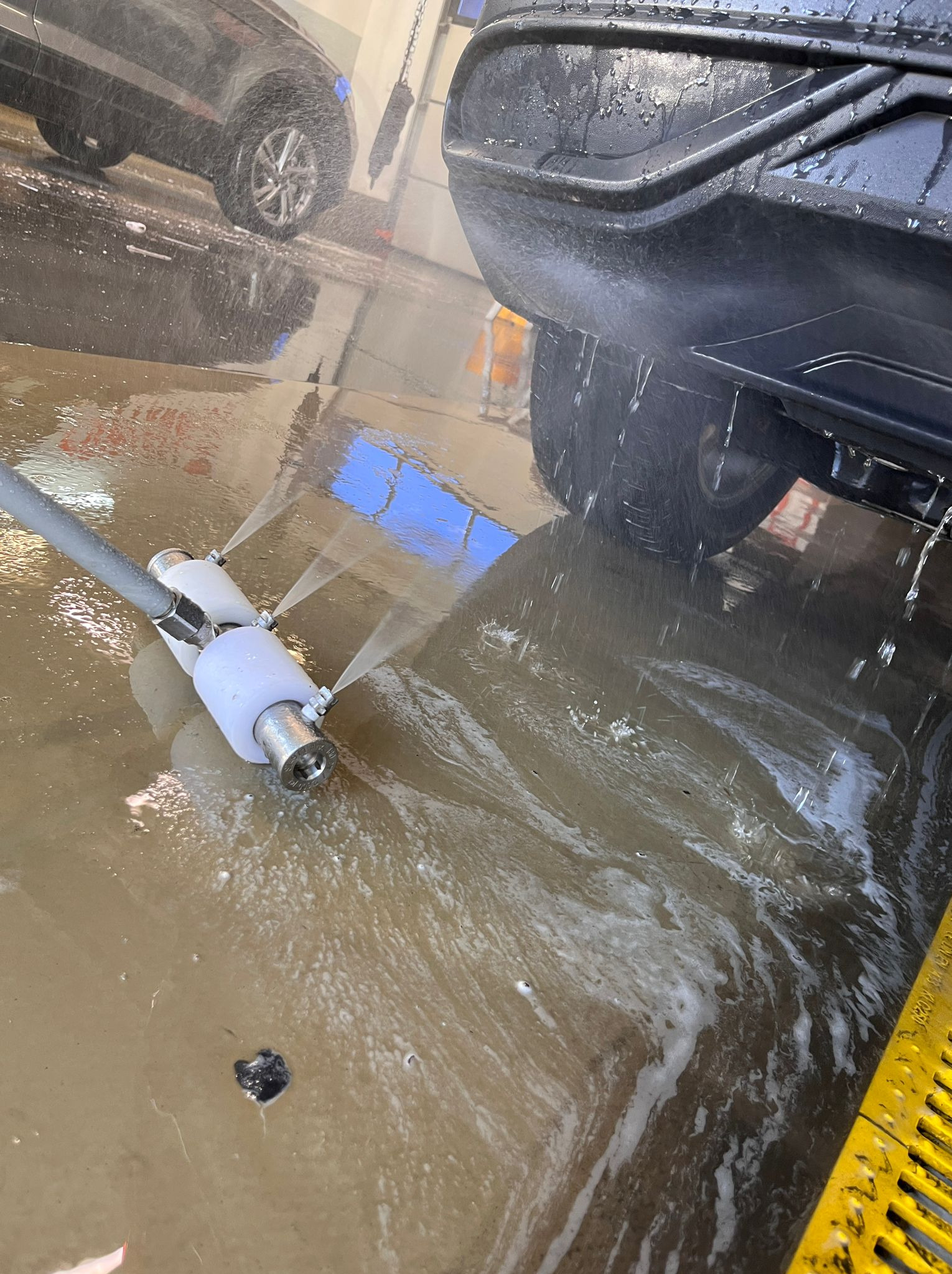 Czarny samochód poddawany myciu podwozia w myjni automatycznej. Woda pod ciśnieniem spryskuje podwozie pojazdu, tworząc efektowną fontannę.