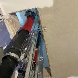 IVAN KASIANCHYK - Montaż pionowych rur wentylacyjnych w otworze stropu, z widocznymi elementami mocującymi i izolacją termiczną na jednej z rur. Prace instalacyjne w surowym wnętrzu.