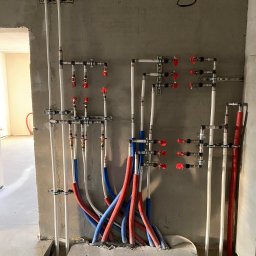 IVAN KASIANCHYK - Węzeł instalacji wodnej w surowym stanie: białe rury pionowe, czerwone i niebieskie poziome, liczne zawory z czerwonymi pokrętłami, szare ściany i podłoga w trakcie remontu.