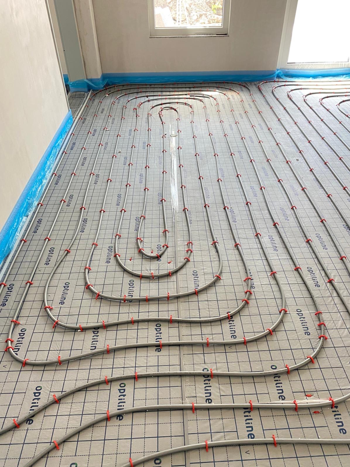 Szare rury ogrzewania podłogowego Optiline, przymocowane czerwonymi klipsami do folii z nadrukiem kratki, ułożone w pętle na podłodze w jasnym pomieszczeniu.