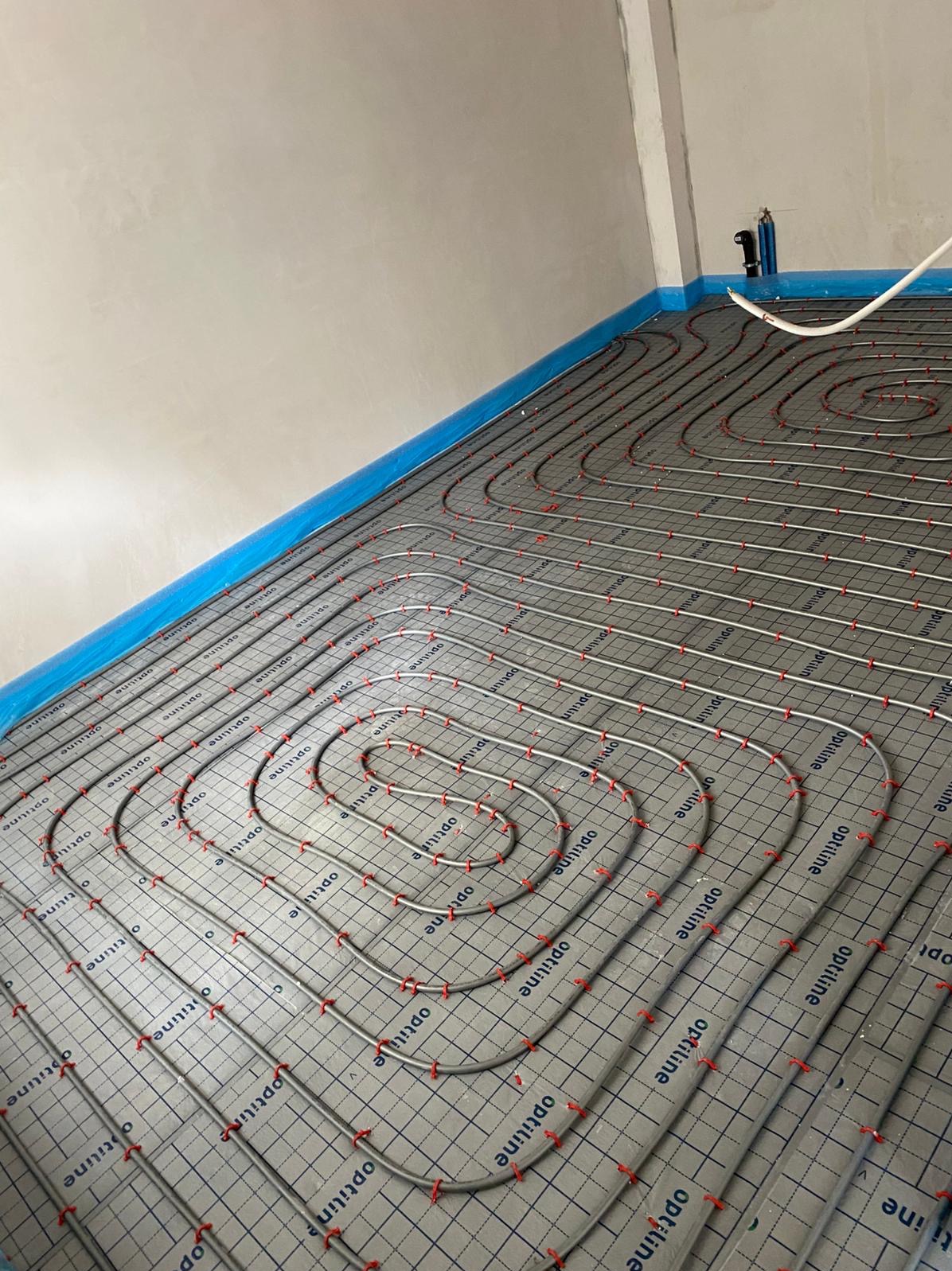 Szare maty grzewcze Optiline z rurkami układane spiralnie na podłodze, zabezpieczone folią przy ścianie. Widok z góry, surowy stan wykończenia.