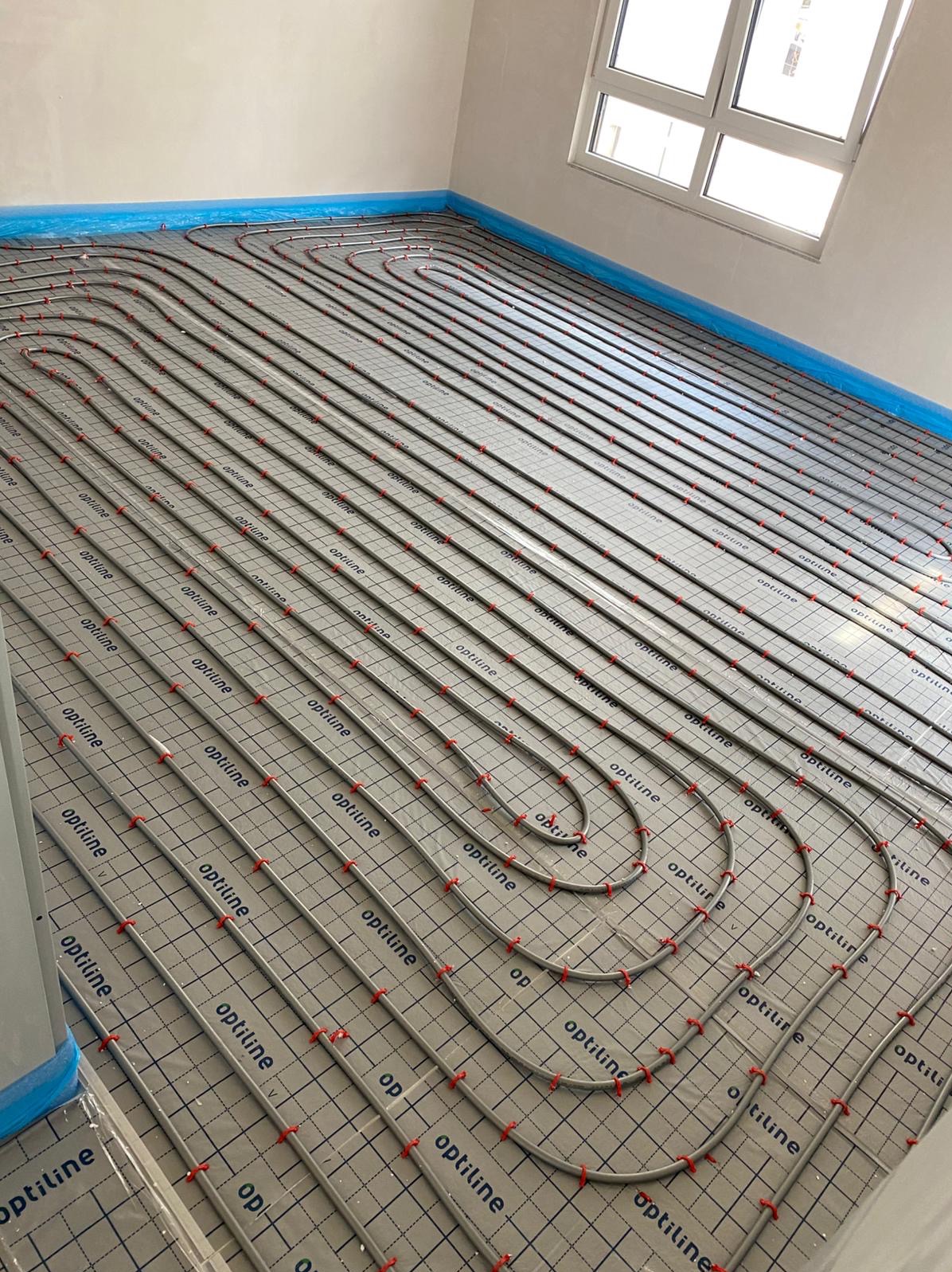 Układanie rur ogrzewania podłogowego na izolacji systemowej z folią i uchwytami mocującymi. Widok na okno i zabezpieczone folią krawędzie ścian.