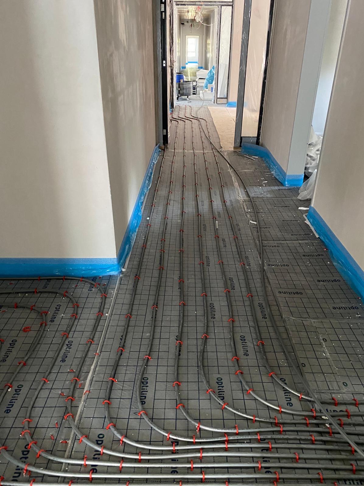 Szara mata pod ogrzewanie podłogowe z rurkami w korytarzu, zabezpieczona niebieską folią przy ścianach. Widok w głąb korytarza, gdzie widać dalszy ciąg instalacji.