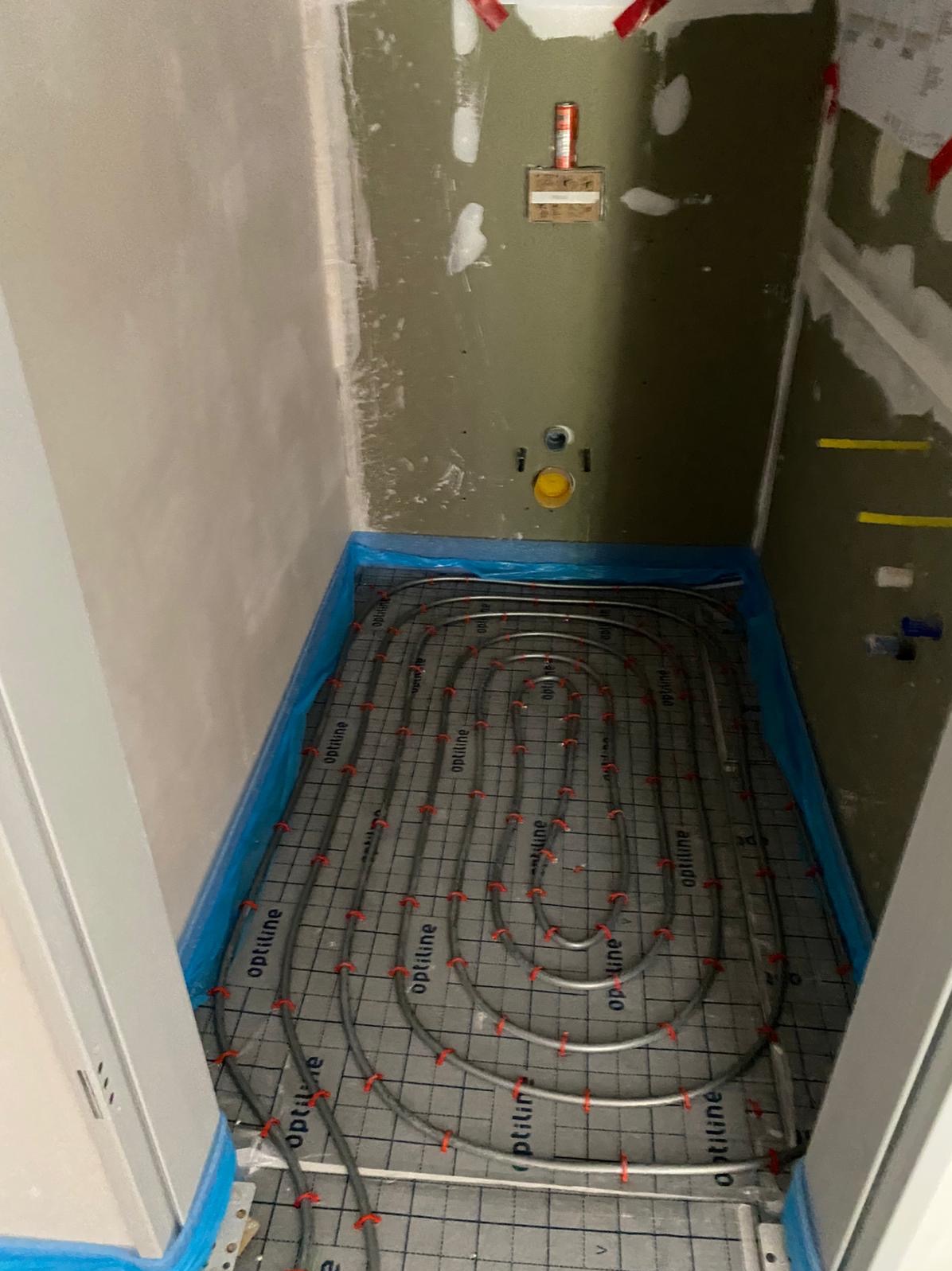 Montaż ogrzewania podłogowego w łazience, widoczne rury ułożone na macie grzewczej, ściany w trakcie przygotowania do wykończenia, zabezpieczone folią.