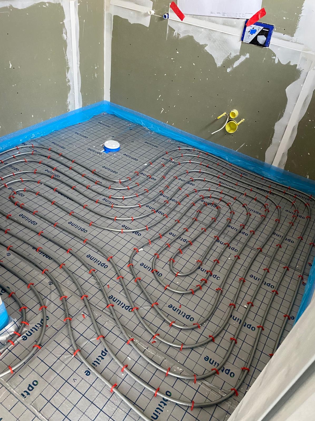 Szare rury ogrzewania podłogowego ułożone na folii z nadrukiem w kratę, mocowane czerwonymi uchwytami, widok z góry na tle surowych ścian w trakcie wykończenia.