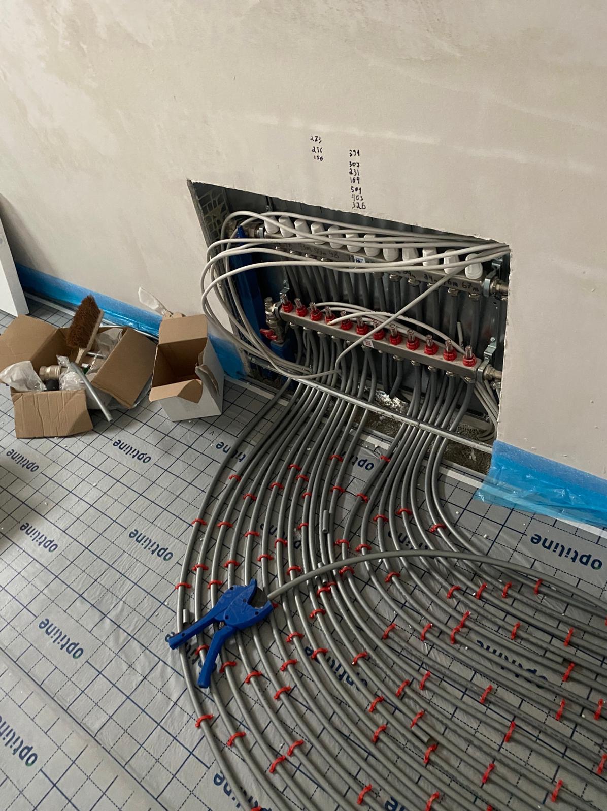 Rozłożona instalacja ogrzewania podłogowego z widocznym rozdzielaczem, rurami mocowanymi do maty izolacyjnej i narzędziami. Ściana z notatkami.