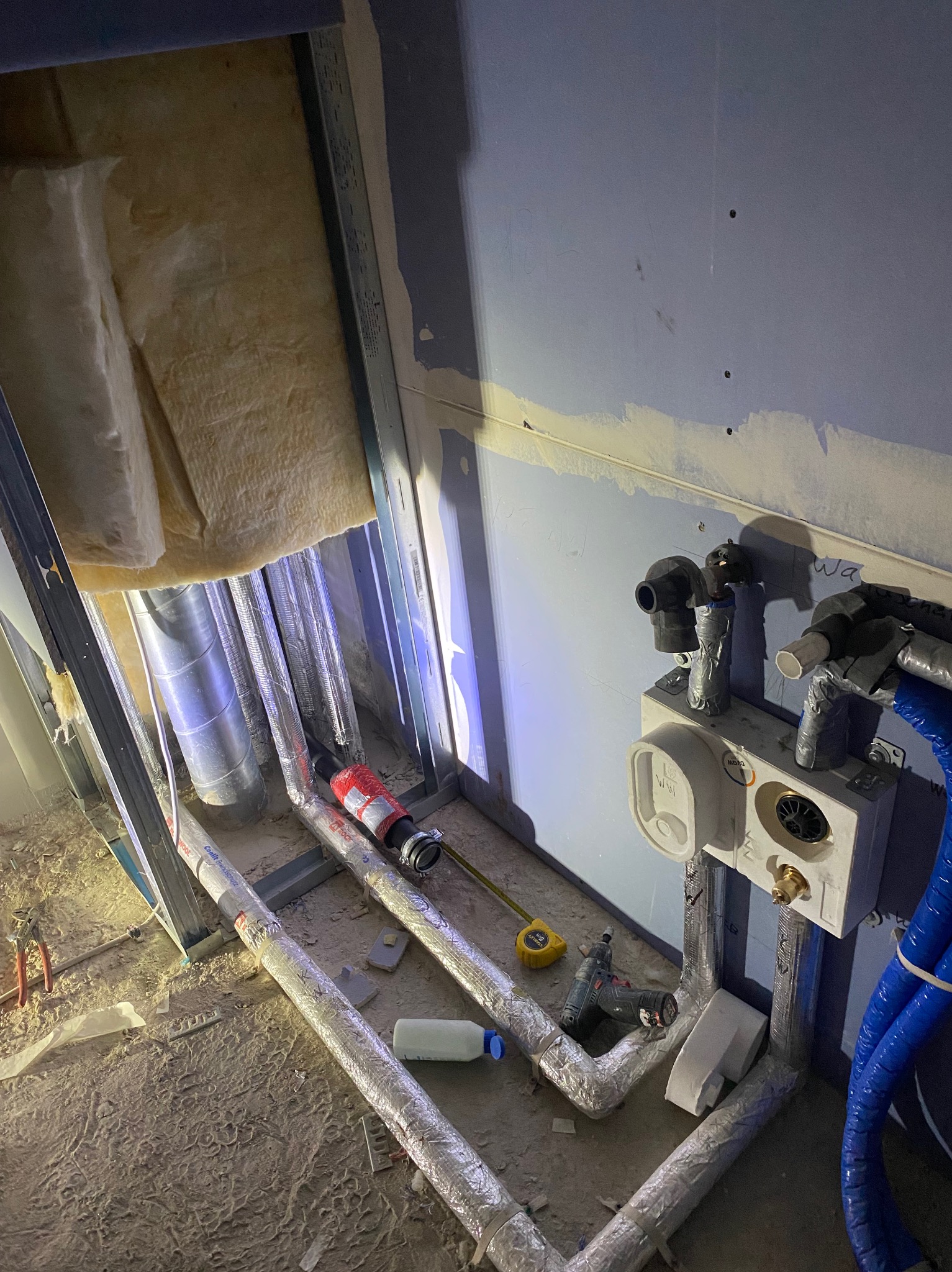 Instalacja wodno-kanalizacyjna w surowym stanie: rury w izolacji, podłączenia do ściany, narzędzia, widoczne elementy konstrukcji. Etap prac przed wykończeniem.