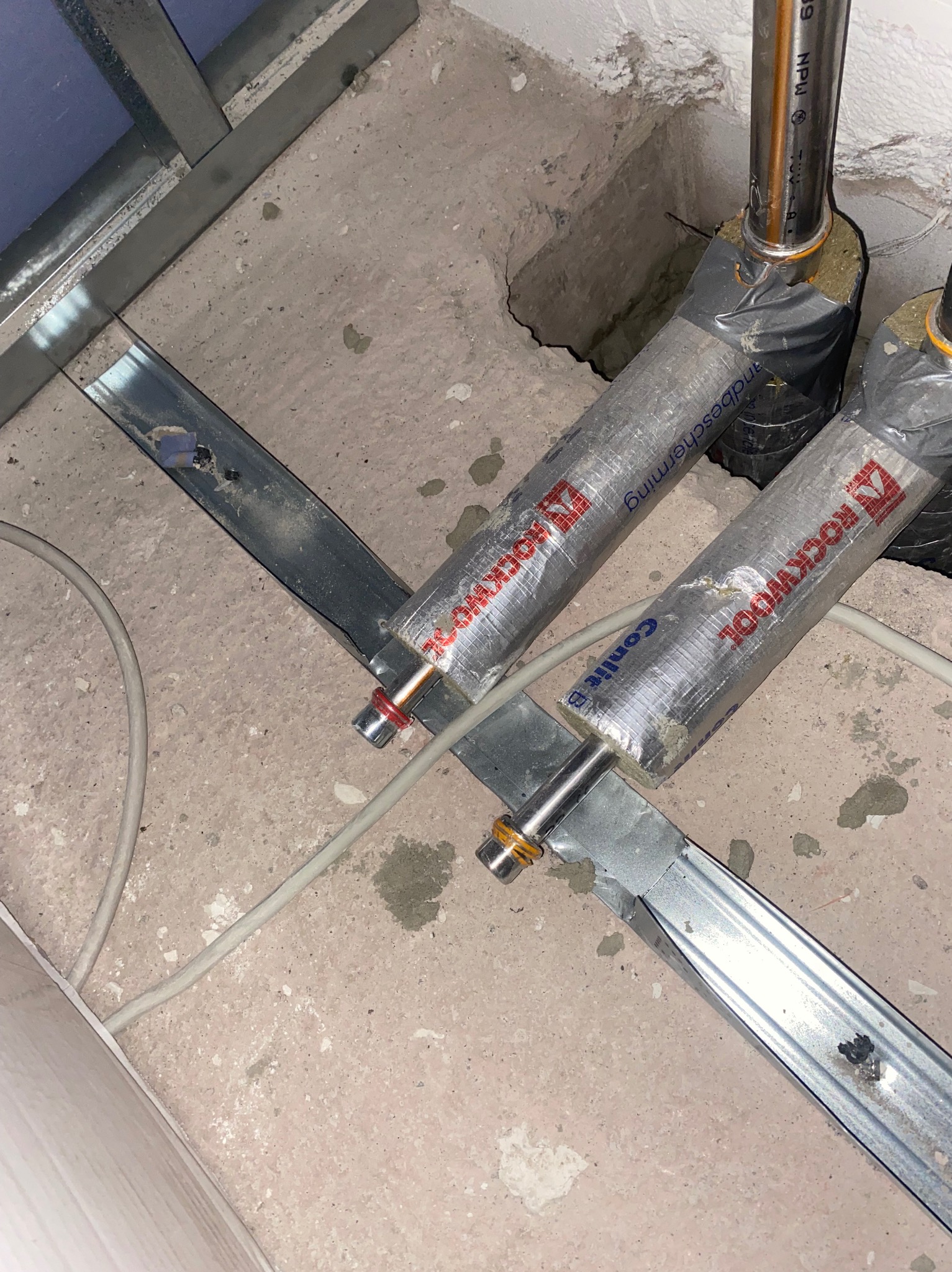 Montaż rur grzewczych z izolacją Rockwool, przymocowanych do metalowych profili w podłodze. Widoczne elementy instalacji i betonowe podłoże.